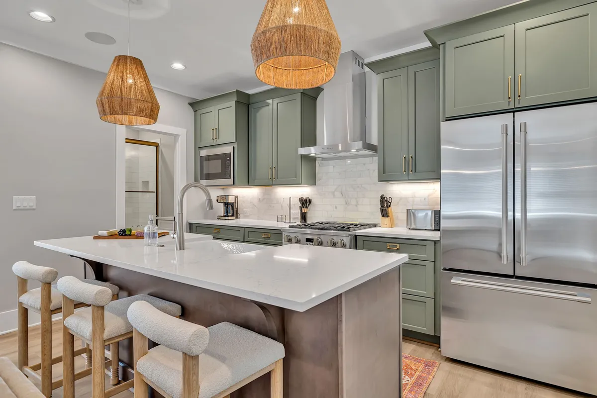 The image showcases a beautifully designed kitchen with a spacious island featuring comfortable seating, stylish green cabinetry, modern pendant lighting, and gleaming stainless steel appliances. The scene suggests a versatile space ideal for cooking, entertaining, and gathering with friends and family, enhanced by an inviting and warm ambiance.