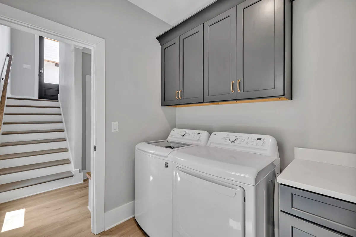 A practical laundry area with washer, dryer, upper cabinets for storage, and stairs leading to another level. The space is neat and organized, suggesting easy, efficient routines for guests.
