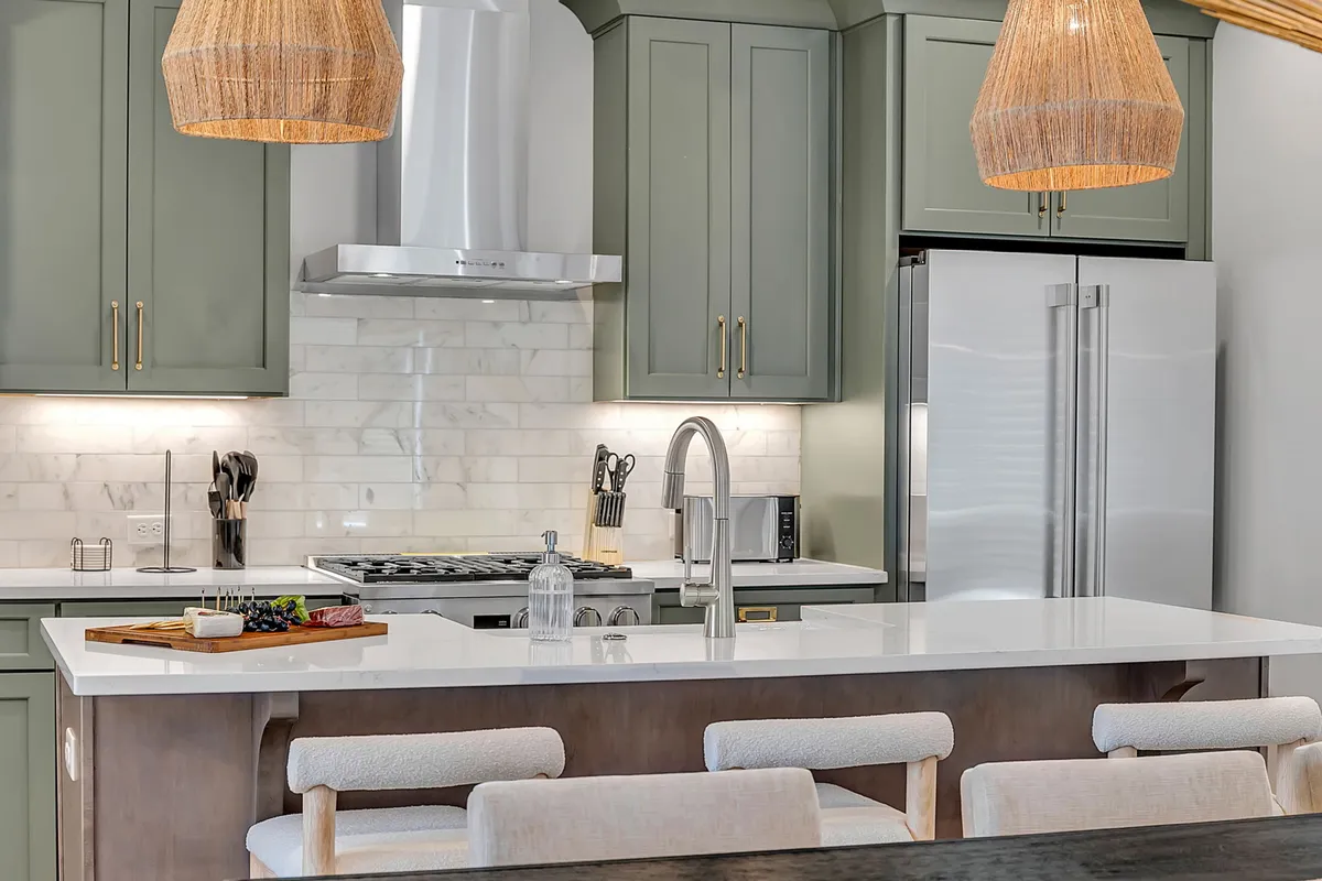 A sleek kitchen featuring a generous island with bar seating, striking hanging light fixtures, stainless steel Monogram appliances, and a marble backsplash. The space is bright, modern, and designed for both cooking and entertaining.