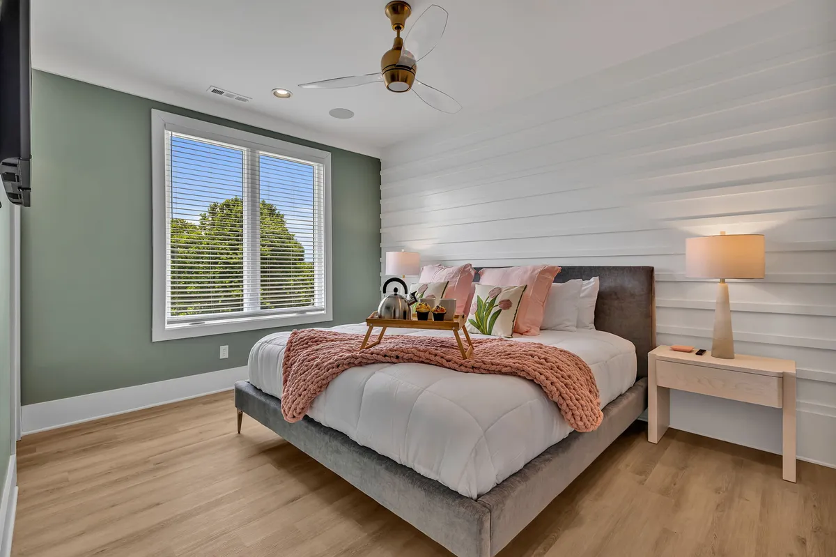 A sunlit bedroom featuring a bed with a pink throw blanket, large windows that let in abundant natural light, and decorative bedside lamps. The space feels cozy and serene, designed for relaxation and comfort.