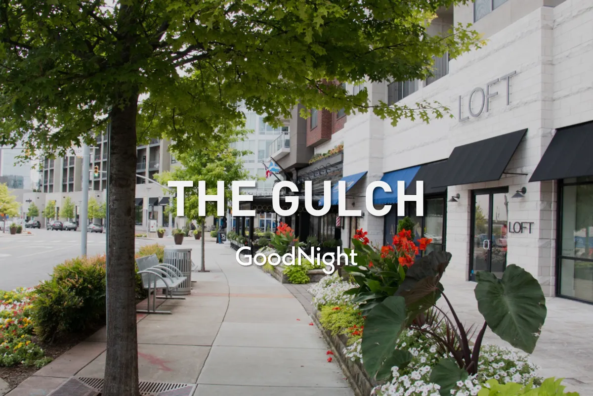 A streetscape showing tree-lined sidewalks, flower beds, and storefronts, with people strolling under the sun. The image conveys a welcoming, walkable neighborhood atmosphere ideal for leisurely walks with pets and spontaneous stops at local shops or cafes.