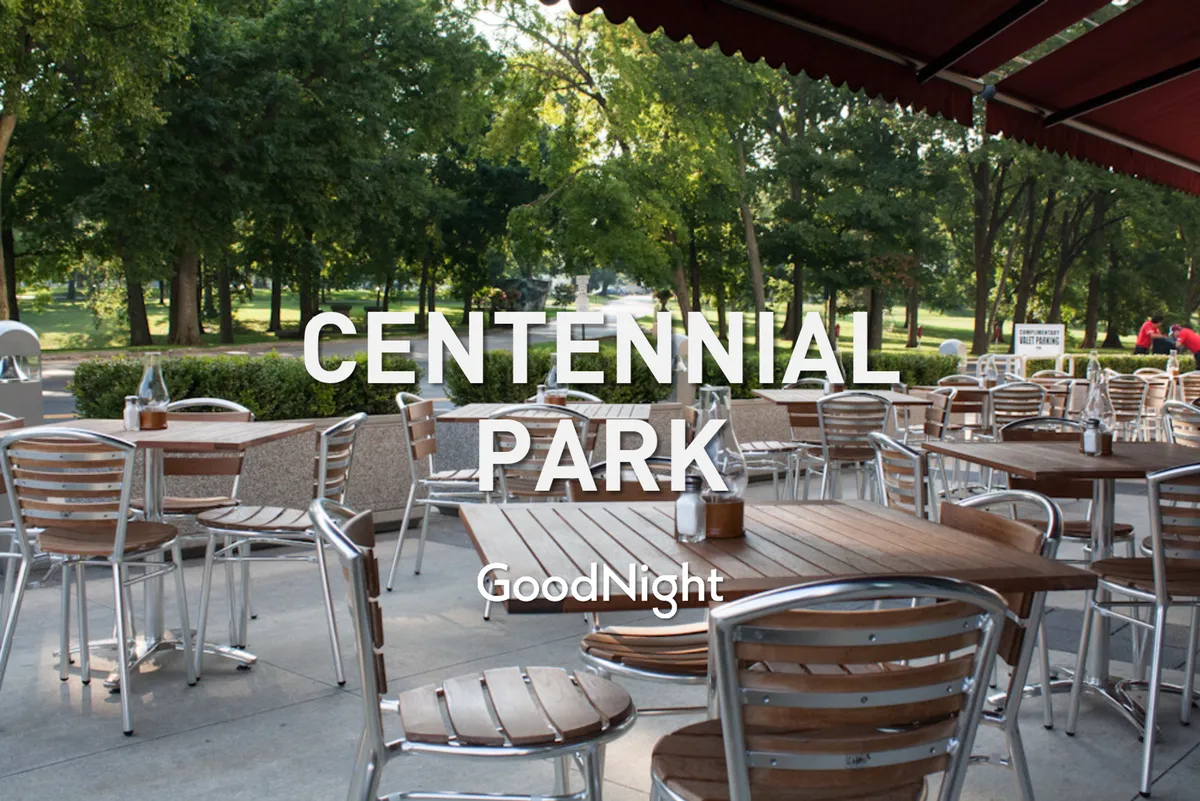 An exterior view of a relaxed outdoor seating area with wooden tables surrounded by lush greenery and a park view. The setup is perfect for dining al fresco, hosting gatherings, or simply people-watching in a natural setting, all activities that are ideal for guests with pets.