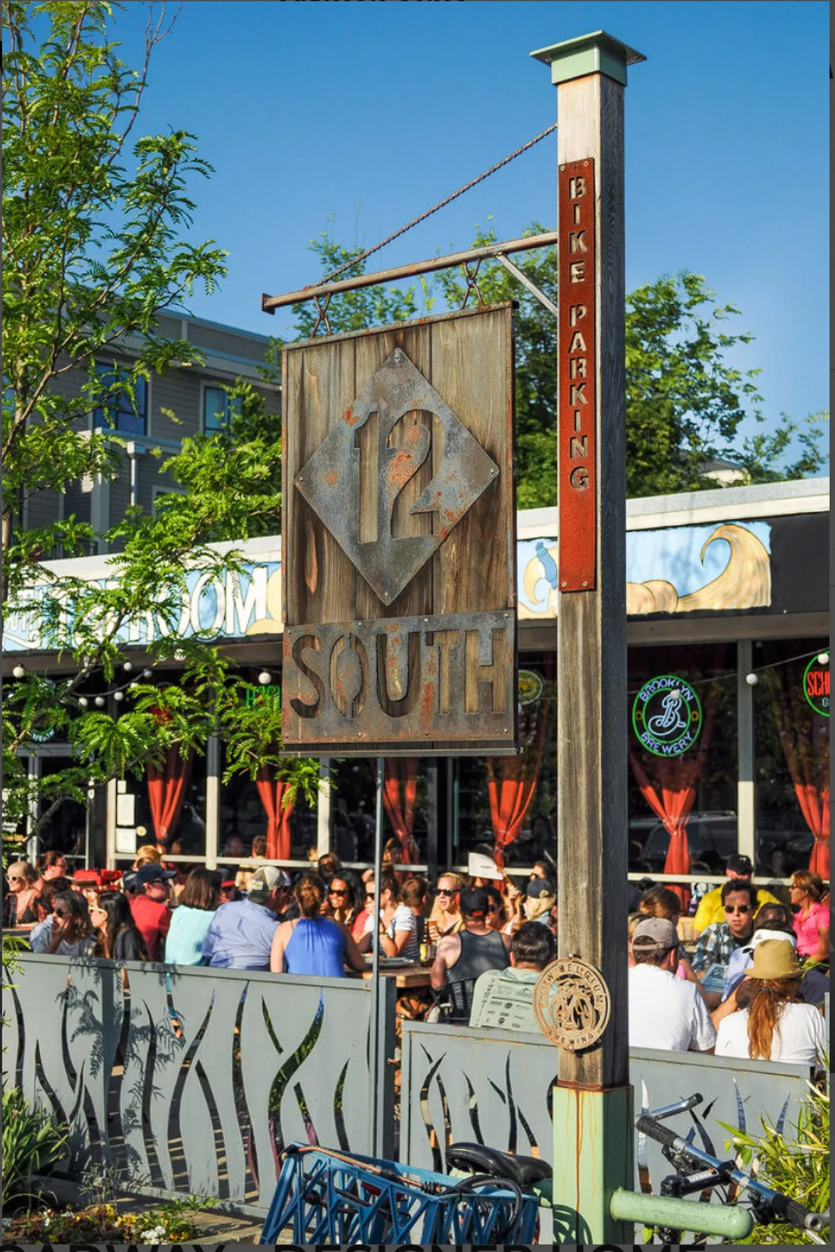 An exterior street scene alive with diners, colorful signage, bike parking, and a festive atmosphere. The decorative fence and visible crowds convey the vibrancy of local eateries and the welcoming feel of the 12 South neighborhood.