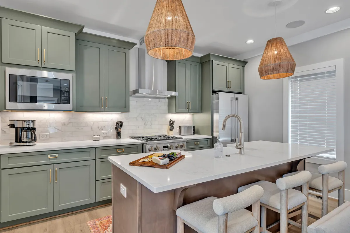 This image showcases a modern kitchen with green cabinetry, a marble countertop, woven pendant lights, and stainless steel appliances. The breakfast bar provides a welcoming spot for casual dining or socializing, and the space is accented by stylish decor, highlighting both functionality and comfort.