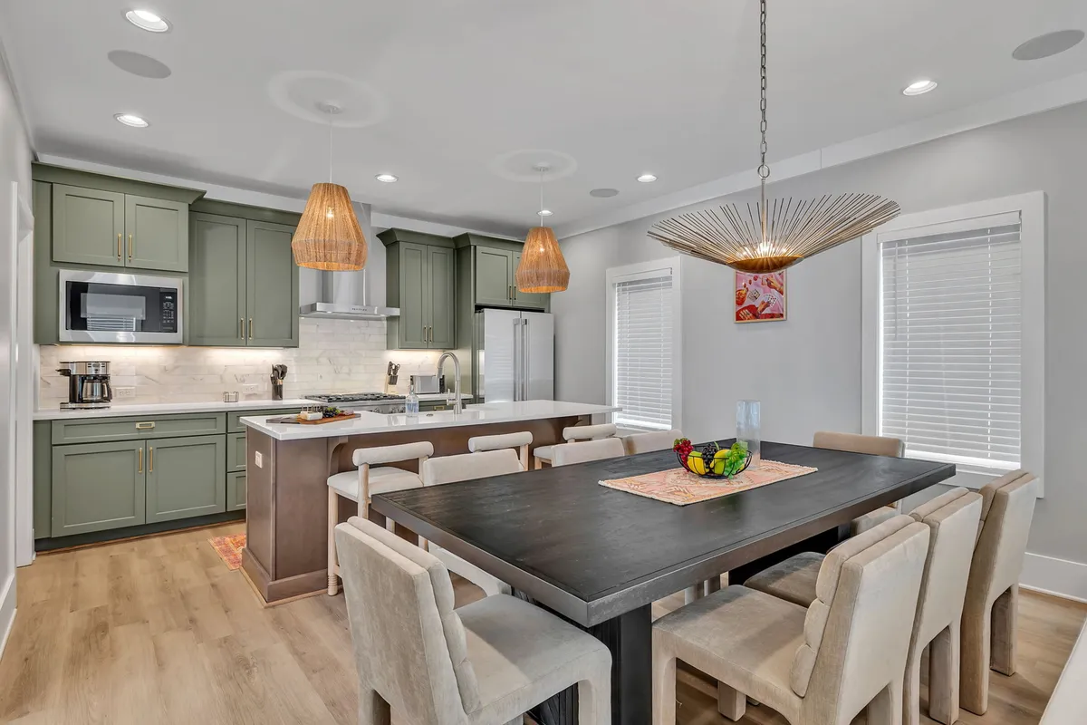 A warm, welcoming view of the kitchen and dining area, with the kitchen island in the foreground, a dining table set for a gathering, and elegant hanging lights creating a cozy ambiance. The scene hints at family meals, entertaining, and shared moments.