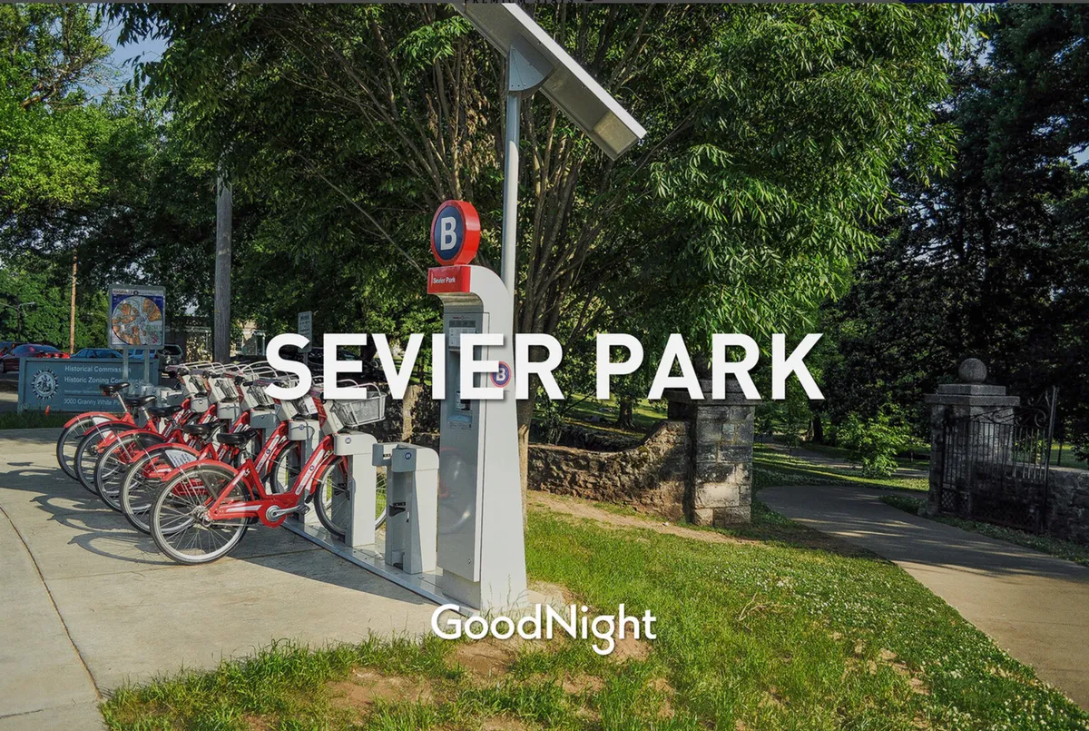 A lively exterior shot featuring a bicycle rental station, park signage, and abundant surrounding greenery. The setting suggests a welcoming public park, ideal for biking, walking, and picnicking. The energetic mood highlights a fun outdoor lifestyle.