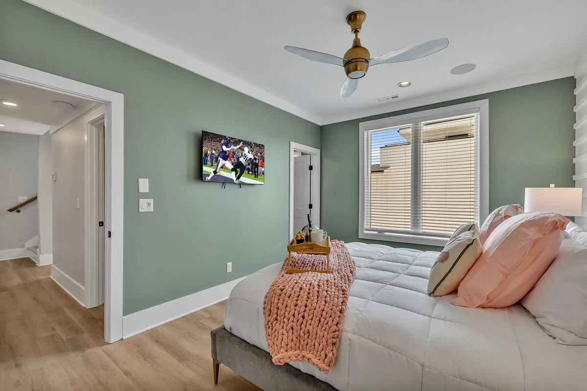 An inviting bedroom with a bed dressed in decorative pillows, a large window flooding the space with natural light, a TV displaying a sports scene, and a colorful throw blanket. The room is cozy and well-appointed for restful stays.