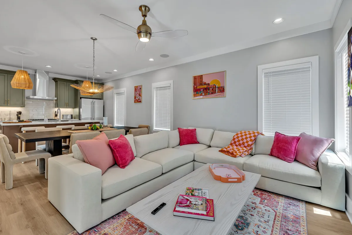A spacious, open-concept living and dining area featuring a large sectional sofa adorned with colorful throw pillows, a wooden dining table that seats eight, vibrant artwork on the walls, and a stylish ceiling fan overhead. The setting is lively and inviting, with hints of both relaxation and social gathering.